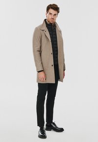 Beżowy wełniany płaszcz z dużym kołnierzem, podwójnym zapięciem i bocznymi kieszeniami, nałożony na czarną kurtkę puchową. Czarne spodnie i buty.