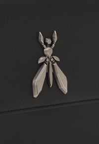 Svart läderprodukt med ett metalliskt emblem i form av ett insekt med skarpa geometriska former och en texturerad yta.