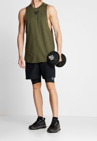 Top sin mangas de color verde olivo con un logo negro, combinado con shorts negros y zapatillas deportivas negras, sosteniendo un dumbbell en la mano derecha.
