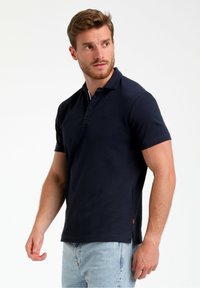 Temno modra polo majica s klasičnim ovratnikom, tri gumbi, kratkimi rokavi, stranskimi razpoki in majhnim logotipom na prsih.