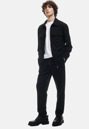 Veste zippée noire avec deux poches poitrine, associée à un pantalon noir ajusté. T-shirt à col rond blanc en dessous. Chaussures noires à lacets.
