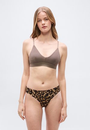 Γυναίκα που φοράει ένα τούπε μπουστάκι και εσώρουχα μπικίνι με λεοπάρ σχέδιο, στέκεται μπροστά σε ένα απλό ανοιχτό γκρι φόντο.