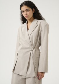 Helle beigen Blazer mit strukturiertem Muster, taillierter Passform und weiten Ärmeln. Verfügt über Seitentaschen und metallische Akzente entlang des Gürtels.