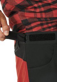 Svarta och röda byxor med kardborrestängning i midjan, som har ett svart strukturerat material och en slät röd panel.