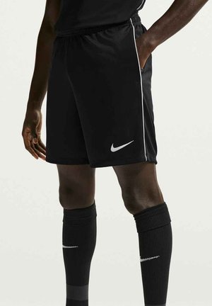 Person trägt schwarze Nike-Sportshorts und lange schwarze Socken, steht mit einer Hand in der Tasche vor einem schlichten Hintergrund.