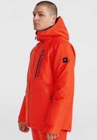 Veste isolée orange vif avec capuche, dotée d'une poche zippée sur la poitrine, de poignets ajustables et d'un écusson logo sur la manche.