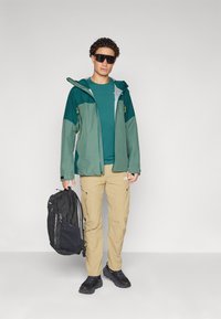 Veste verte résistante à l'eau avec des accents gris, accompagnée d'une chemise teal et d'un pantalon cargo beige. Un sac à dos noir et des chaussures de sport complètent l'ensemble.