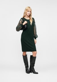 Μακρυμάνικο, σκούρο πράσινο φόρεμα με διαφανείς, κεντημένες μανίκες; συνδυασμένο με μαύρες μπότες μέχρι το γόνατο και μια μικρή μαύρη τσάντα.