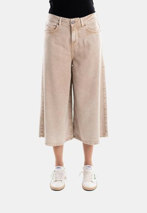 Pantalon beige large et raccourci porté avec des baskets blanches, modèle debout sur un fond uni.