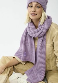 Lila gestrickter Schal mit gerippter Textur, um den Hals drapiert, mit einem kleinen Logodetail, kombiniert mit einer passenden Mütze.