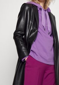 Abrigo de cuero sintético negro sobre una sudadera morada con cordones y una camiseta morada, combinado con pantalones morados oscuros. Se aprecian texturas suaves.