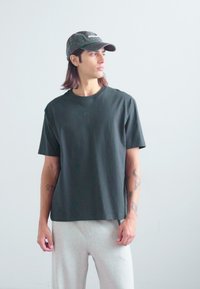 Tricou scurt cu maneci scurte, de culoare verde închis, cu guler rotund și un logo subtil. Este purtat cu pantaloni de trening gri deschis și o șapcă cu model.