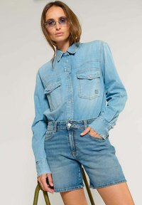 Lichtblauwe denim overhemd met knoopsluiting en twee borstzakken, gecombineerd met lichtblauwe denim shorts. Soepele textuur, casual pasvorm en contrasterende stiksels.
