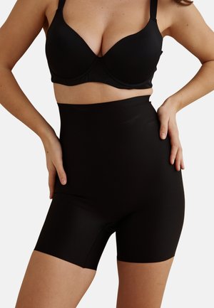 Femme portant un short gainant noir taille haute et un soutien-gorge noir, mains posées sur les hanches, torse et haut des jambes visibles.