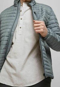 Muž v světle šedé prošívané bundě přes béžový henley svetr, drží bundu otevřenou a ukazuje knoflíky a texturu látky svetry.