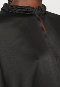 Gros plan sur un chemisier noir texturé avec un col tressé et une encolure fendue à l'avant sur une peau foncée.