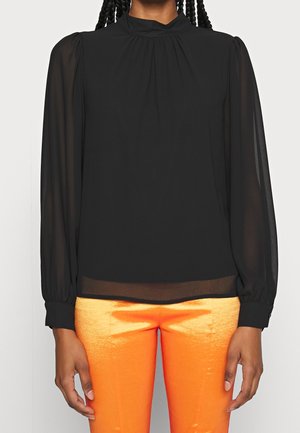 Person iført en langærmet sort gennemsigtig bluse med høj hals og skinnende, lyse orange bukser, stående foran en ensfarvet baggrund.