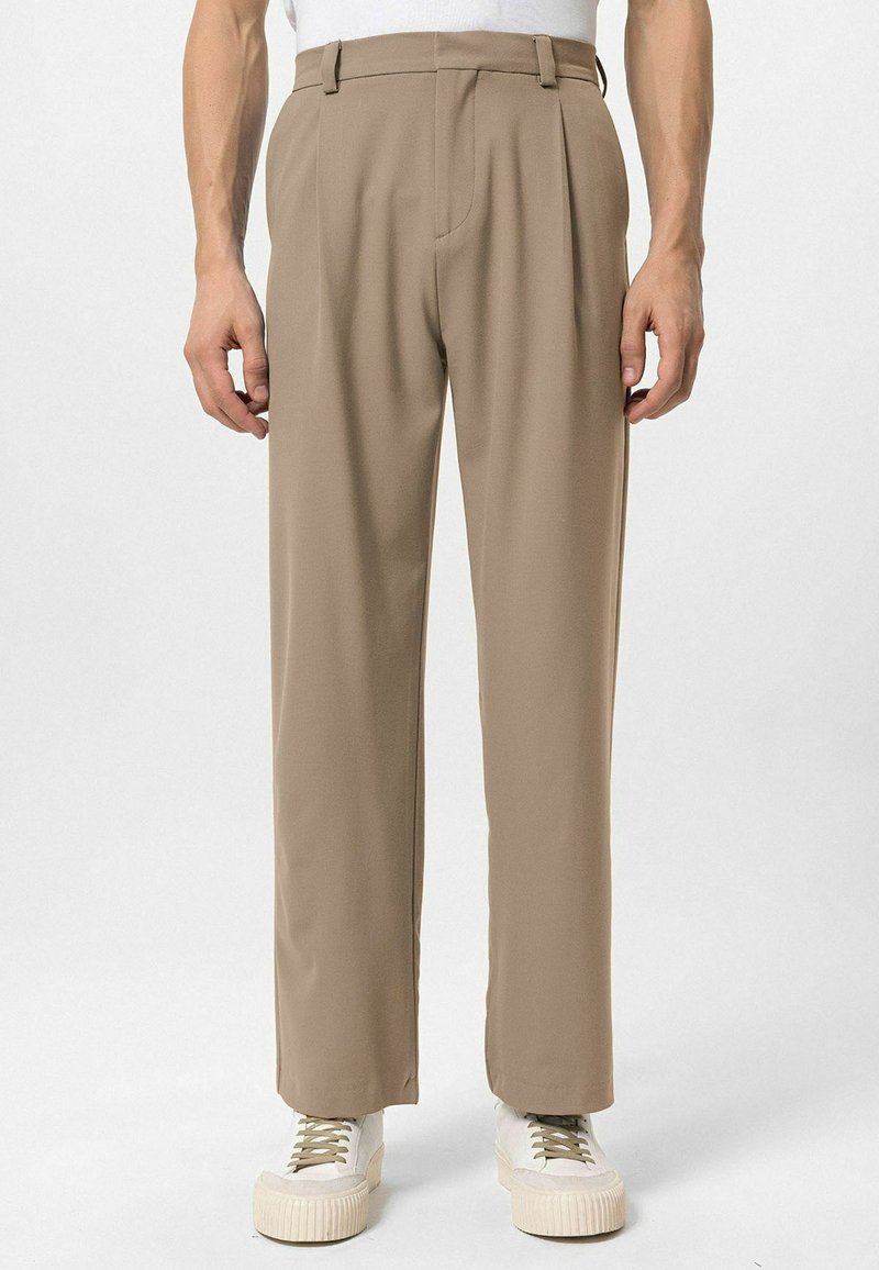 Beige broek met een getailleerde pasvorm, geplooide voorkant en zijzakken. De stof lijkt glad, gecombineerd met lichtgekleurde sneakers.