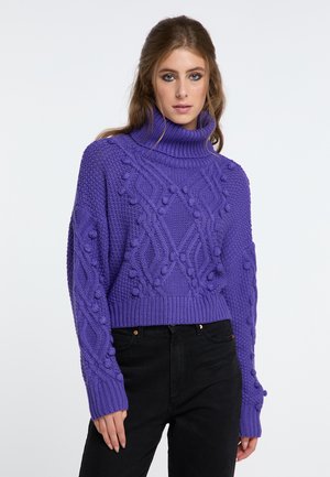 Frau trägt einen violetten grob gestrickten Rollkragenpullover mit strukturierten Zopf- und Noppenmustern, kombiniert mit schwarzen hoch taillierten Hosen.
