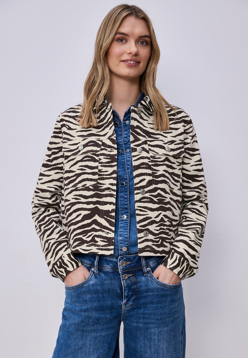 Ung kvinde iført en beige og sort zebra-print jakke over en blå denimskjorte og jeans, hænderne i lommerne, neutral baggrund.