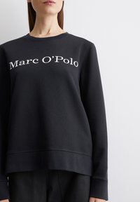 Must muste, mille on ümar krae, pikad varrukad ja madalam lõhk. Esiküljel on valge "Marc O'Polo" tekstitrükk. Sile tekstuur.