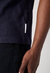 Marineblaue Baumwoll-T-Shirt mit feiner Textur, ausgestattet mit einem seitlichen Etikett und kurzen Ärmeln, kombiniert mit dunklen Jeans.