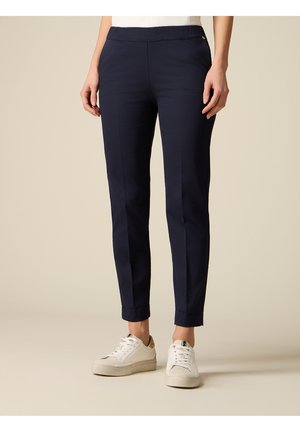 Pantalon marine slim ajusté longueur cheville porté avec des baskets blanches, vue de face de la taille aux pieds sur fond beige.