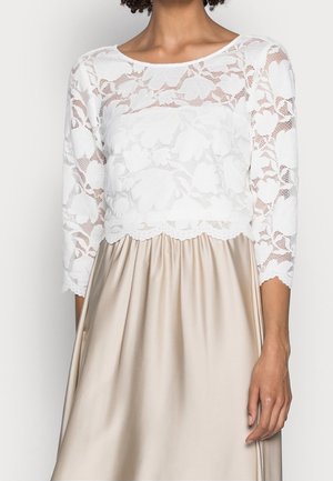 Haut en dentelle florale blanche à manches trois-quarts, associé à une jupe en satin beige, porté par une personne du cou jusqu'à la mi-cuisse.