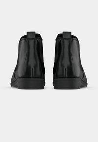 O pereche de cizme Chelsea din piele neagră, văzute din spate, cu panouri laterale elastice și tab-uri pentru tragere, pe un fundal alb simplu.