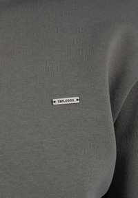 Tejido de sudadera gris con costillas y una etiqueta metálica. La etiqueta presenta "SMILODOX" en una fuente negrita, ubicada en la zona del pecho.