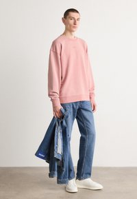 Sudadera rosa claro con cuello redondo, corte relajado y puños acanalados. Combinada con jeans de mezclilla azules y zapatillas blancas, sosteniendo una chaqueta de mezclilla.