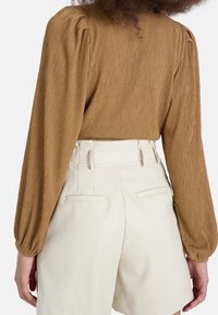 Maglietta lunga a maniche con pieghe di colore marrone abbinata a pantaloni corti di colore crema; la maglietta presenta maniche a sbuffo e un tessuto testurizzato. I pantaloni hanno tasche laterali.