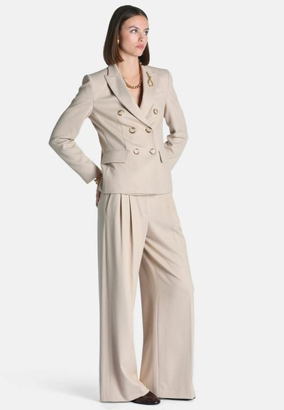 Femme aux cheveux bruns raides portant un blazer beige croisé et un pantalon à jambes larges, accessoirisée de bijoux dorés, debout les mains derrière.