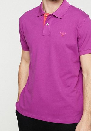 Man wearing a short-sleeve purple polo shirt with an orange button placket and GANT logo on the left chest.