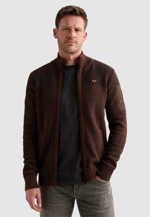 Homme aux cheveux bruns courts porte un pull zippé marron foncé texturé sur une chemise noire et un jean gris, debout avec une main dans la poche.