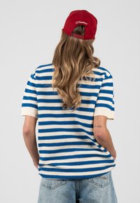 Persona con cabello largo y ondulado lleva una gorra roja, camiseta de manga corta a rayas azul y blanca, y vaqueros vaqueros azul claro, mirando de espaldas a la cámara.