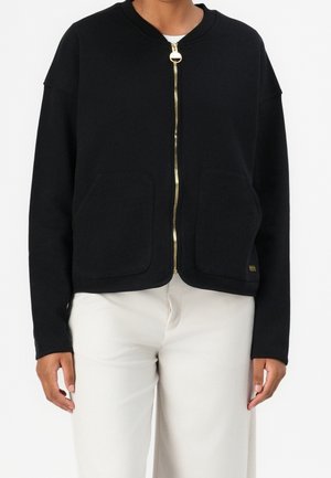 Personne portant une veste noire zippée avec des poches avant et une fermeture éclair dorée, associée à un pantalon blanc, debout, bras détendus.