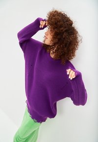 Maglione viola lavorato a maglia con maniche lunghe, caratterizzato da una trama a coste, spacchi laterali e un orlo a onde, abbinato a pantaloni verdi in tessuto testurizzato.
