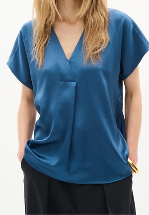 Kobieta ubrana w niebieską bluzkę o teksturze, z krótkim rękawem i dekoltem w serek, czarne spodnie oraz złotą bransoletkę na lewej ręce.