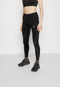 Schwarze Sportleggings mit glatter Textur, ausgestattet mit Netzeinsätzen und weißen seitlichen Streifen. Kombiniert mit einem schwarzen Sport-BH und schwarzen Turnschuhen.