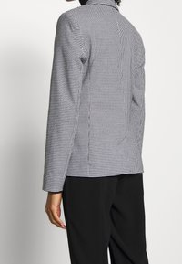 Blazer s černobílým vzorem houndstooth, přiléhavého střihu, s klopovým límcem a dlouhými rukávy, zobrazený zezadu.