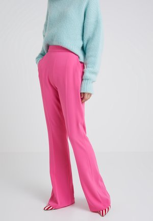 Persoon die felroze rechte broek draagt, een lichtblauwe pluizige trui en rood-wit gestreepte puntige schoenen tegen een effen achtergrond.