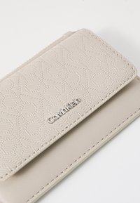 Calvin Klein lommebok i lys beige teksturert skinn med preget logo. Har en jevn overflate og stikninger i kantene, med glidelås.