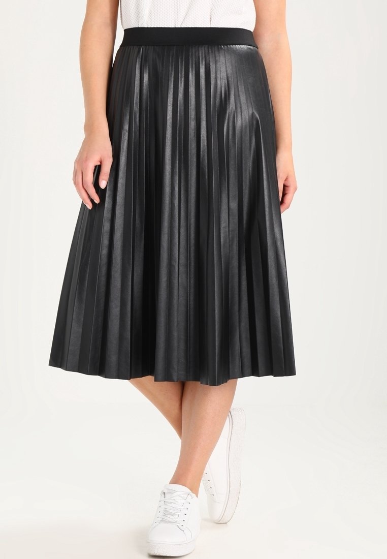 Femme portant une jupe midi plissée noire avec une taille cintrée, associée à des baskets blanches et un haut blanc, debout, jambes croisées.