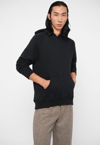 Felpa nera con tasca a marsupio e polsini a coste. Il tessuto appare morbido e il design presenta una vestibilità ampia con cappuccio regolabile.