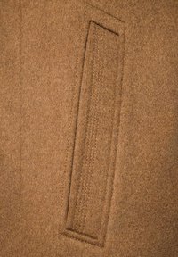 Brązowa wełniana tkanina o gładkiej fakturze z prostokątną kieszenią podkreśloną kontrastującym ściegiem. Szczegół podkreśla strukturę tkaniny.