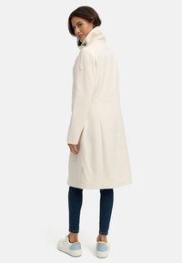 Abrigo blanco de mezcla de lana con cuello alto, aberturas laterales y detalle de logo en la manga, combinado con jeans oscuros y zapatillas blancas con acentos azules.