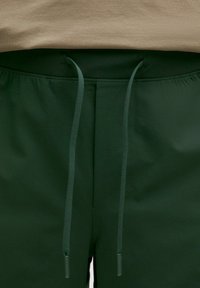 Mörkgröna shorts med dragsko i midjan. Slät textur och elastiskt midjeband, med två tunna snören med platta metallspetsar.