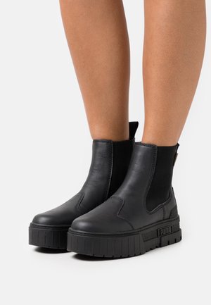 Zwarte leren enkellaarsjes met dikke plateauzolen gedragen op blote benen tegen een effen witte achtergrond.