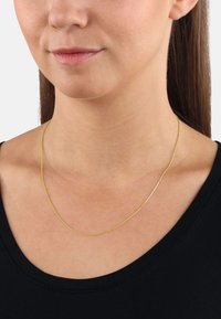 Collar de cadena de color dorado con un fino diseño retorcido, apoyado sobre un cuello desnudo, llevado sobre una blusa negra.